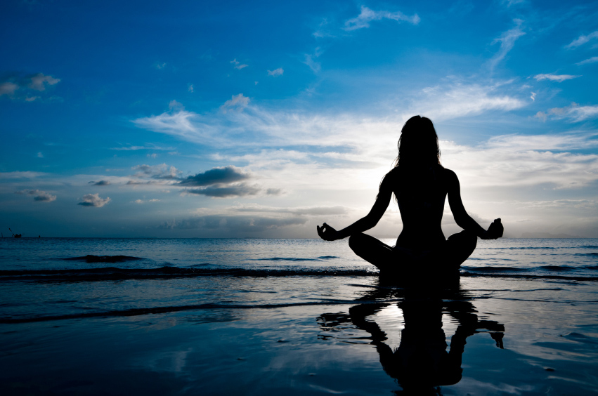 woman sat in sukhasana yoga pose practising mediation sat with her reflection on the wet sand